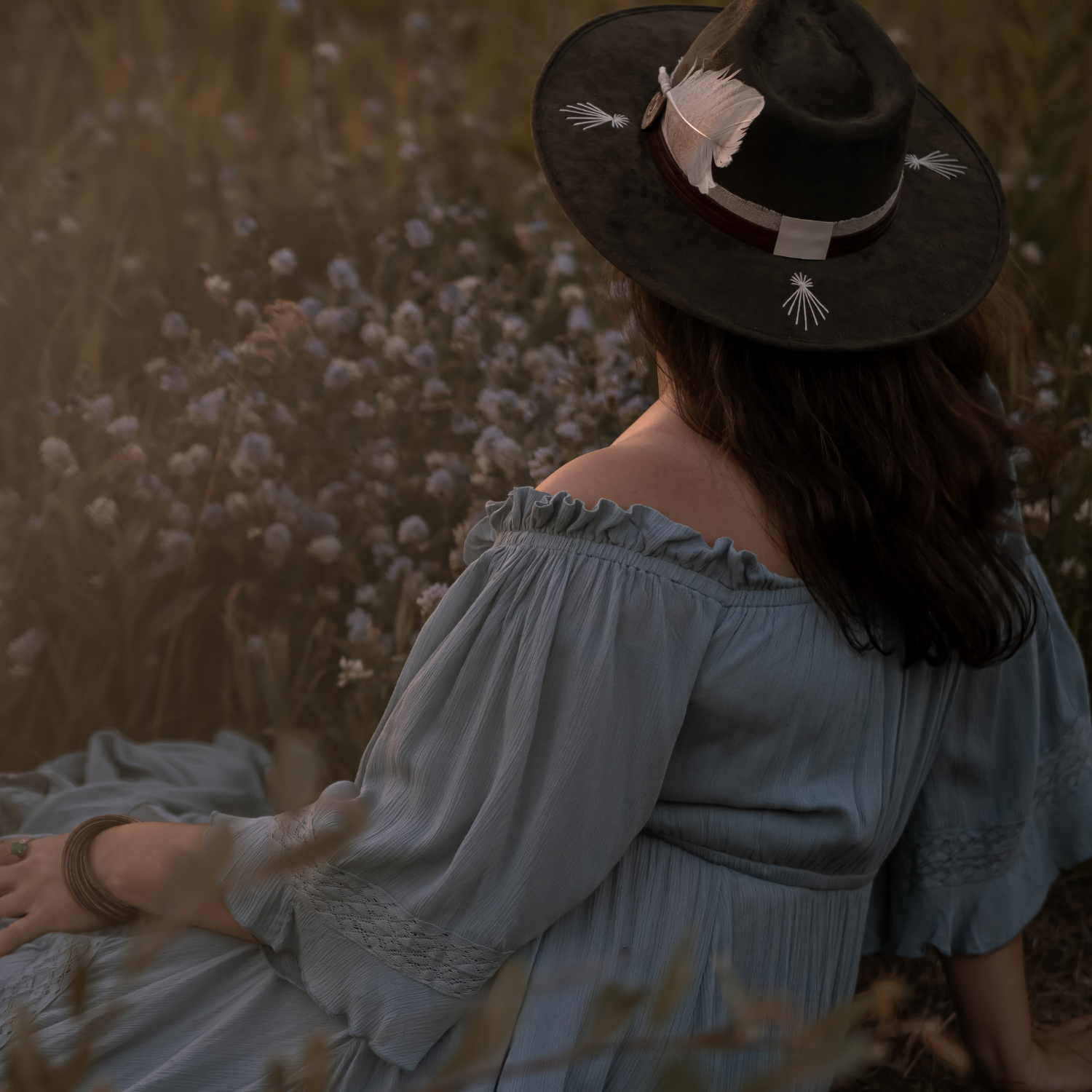 Woman wearing a forest green Kindred fedora hat and periwinkle dress seated with her back to the viewer in a field of periwinkle flowers.