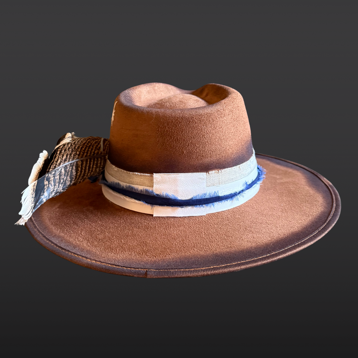 A tobacco wide-brimmed fedora shown from the back, featuring a hand-torched textured finish, cream leather band with navy cotton wrap, and turkey feather accent.