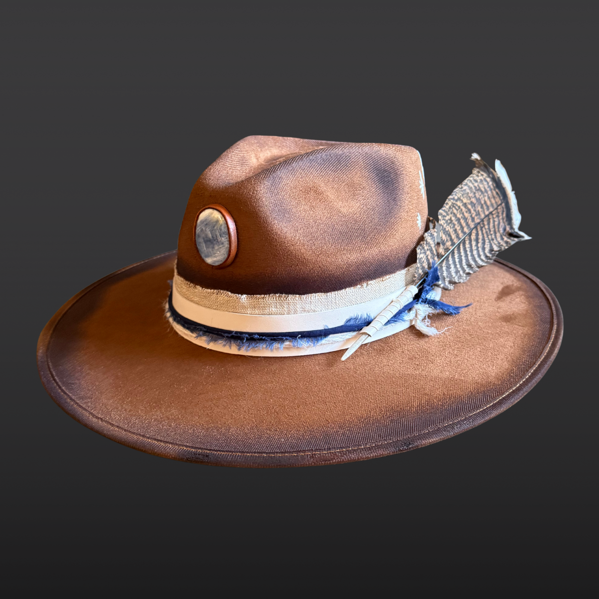 Main view: A tobacco wide-brimmed fedora featuring an all-over hand-torched finish, a cream leather band with navy cotton wrap, a blue kyanite stone, an embroidered sunburst, and a turkey feather accent.