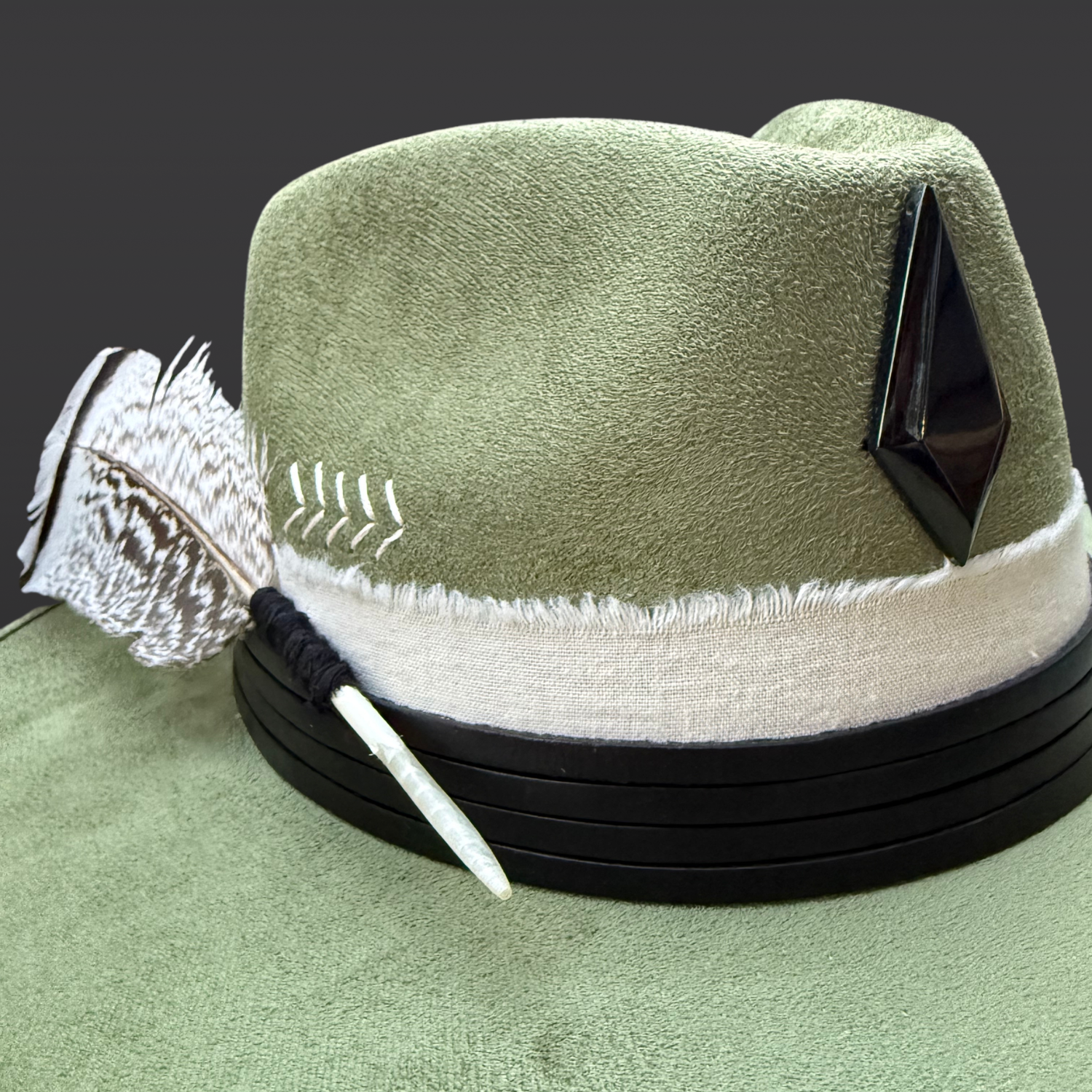 One of a kind sage green artisan fedora hat with obsidian stone, black leather band, white cotton wrap, feather detail, and hand embroidered directional symbols, close up of details by Inara Hat Co.