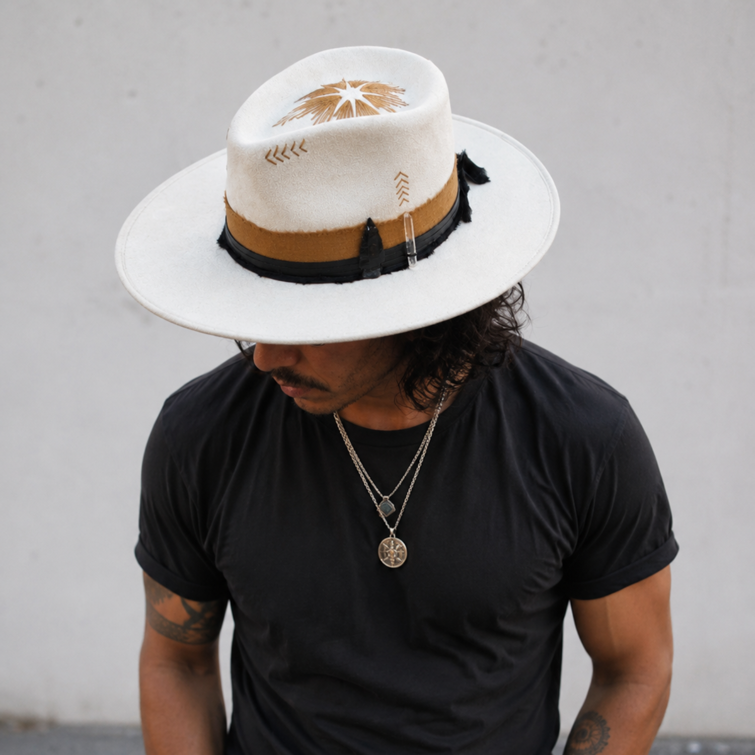 Man in his late 30s wearing the Sunfire Ascend wide-brim cream vegan suede hat with a tall teardrop crown, flat structured brim, tan leather band, black trim, clear quartz crystal, black crystal, and decorative stitching positioned on the left side. Styled with fitted black streetwear and subtle boho accessories, his head is tilted down to showcase the handcrafted details against a clean urban background.
