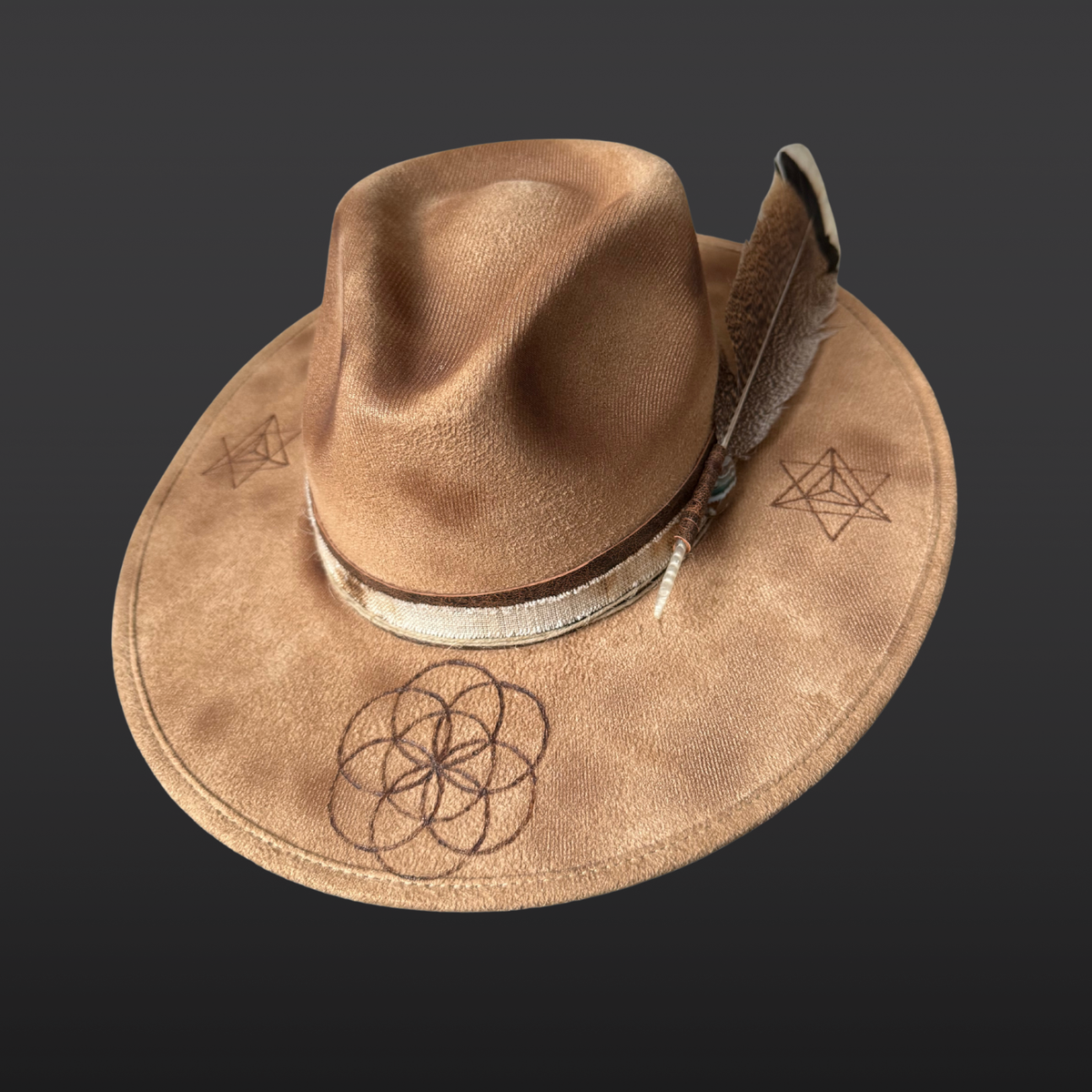 Front angle:
A wide-brimmed camel-colored fedora featuring hand-burned Seed of Life and Merkaba geometric designs on the crown, with a dark brown leather band and turkey feather accent.