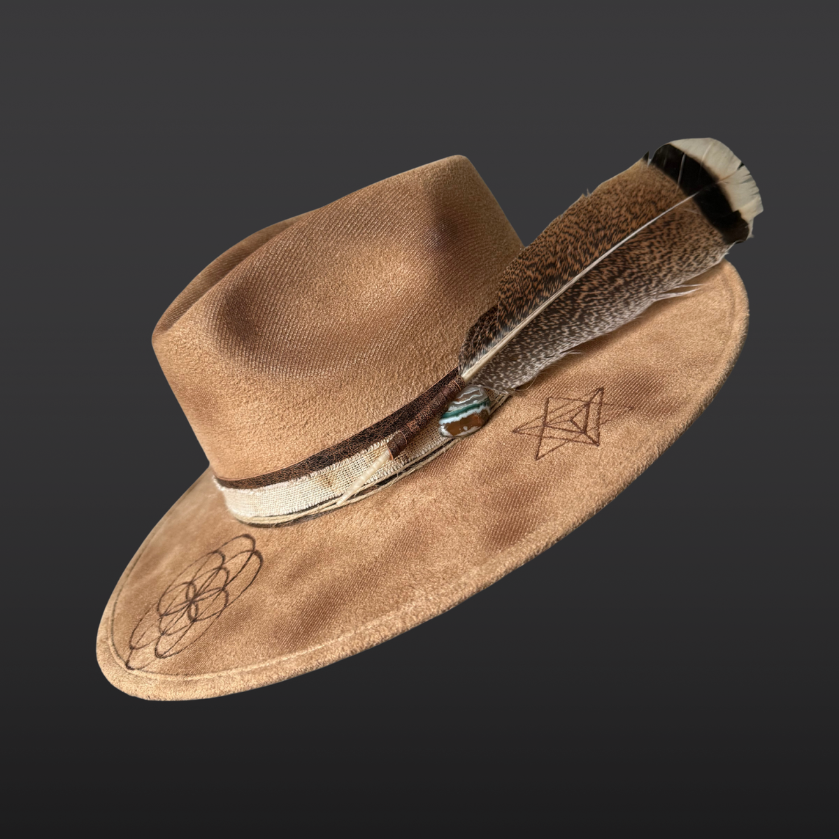 Main view:
A wide-brimmed camel-colored fedora featuring a dark brown leather band, a turkey feather accent, an agate stone, and hand-burned Seed of Life and Merkaba geometric designs on the crown.