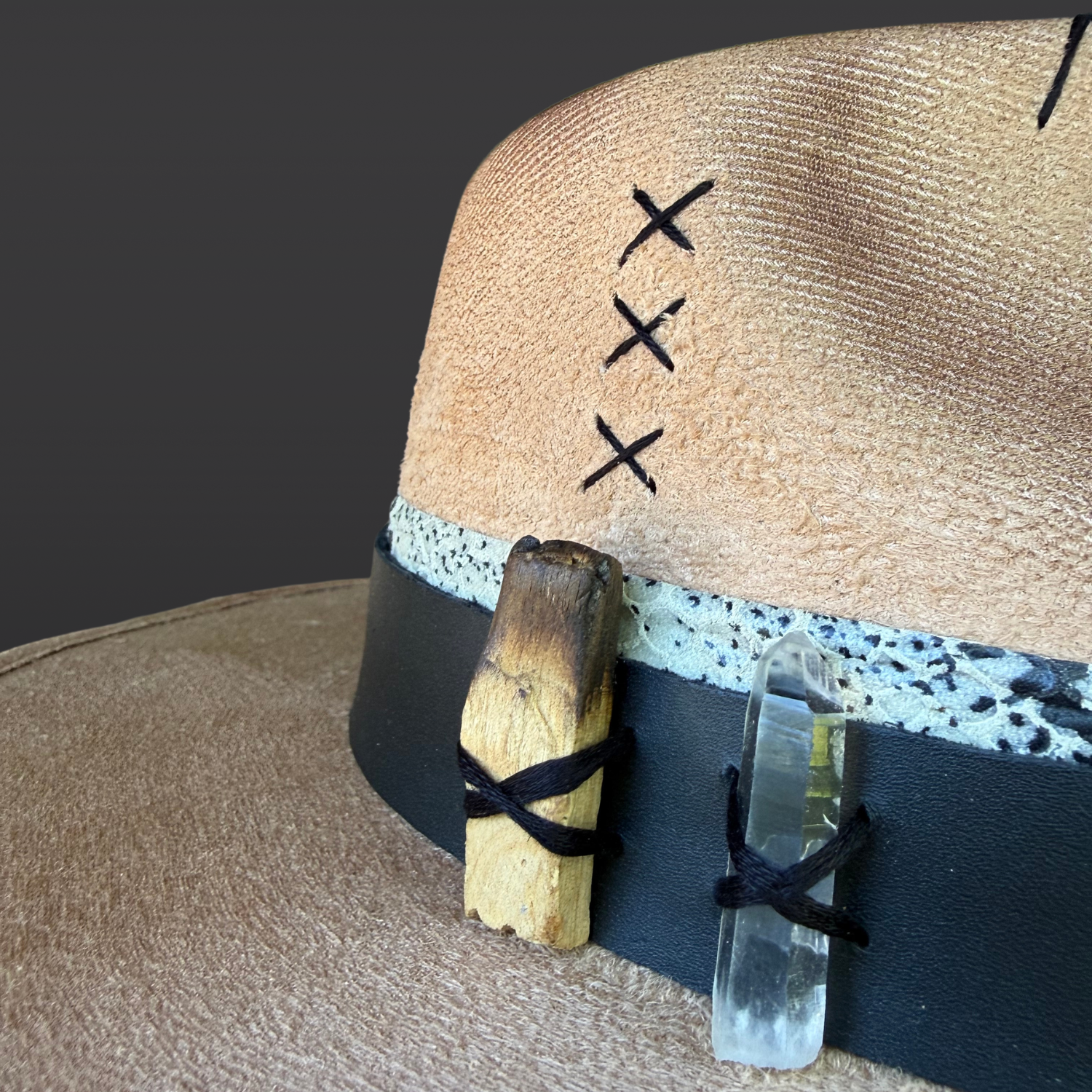 Close-up of clear quartz crystal, naturally burnt palo santo, and turkey feather attached to the side of a camel wide brim fedora.