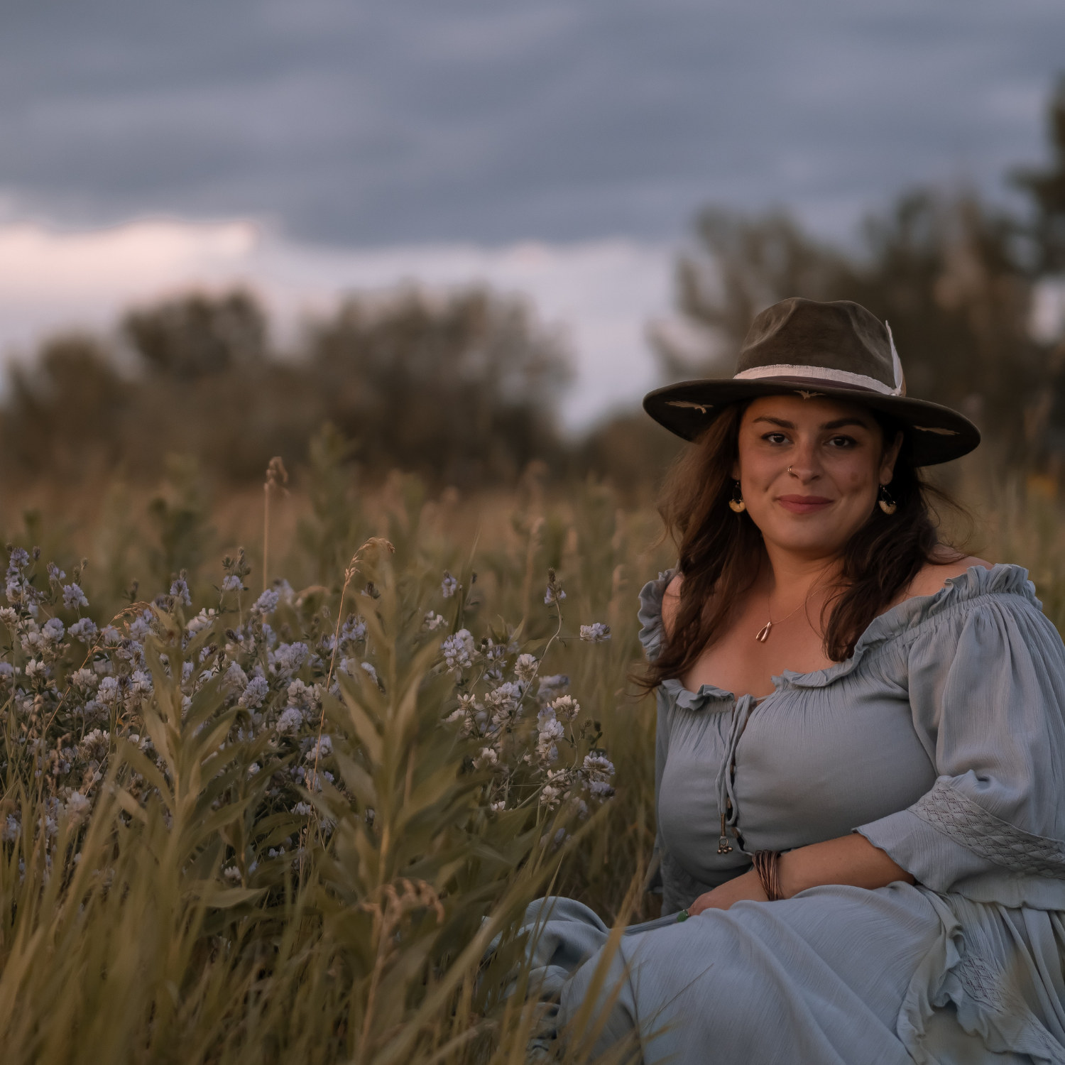 Woman wearing a forest green Kindred fedora hat and periwinkle dress seated in a field of periwinkle flowers beneath a moody sky