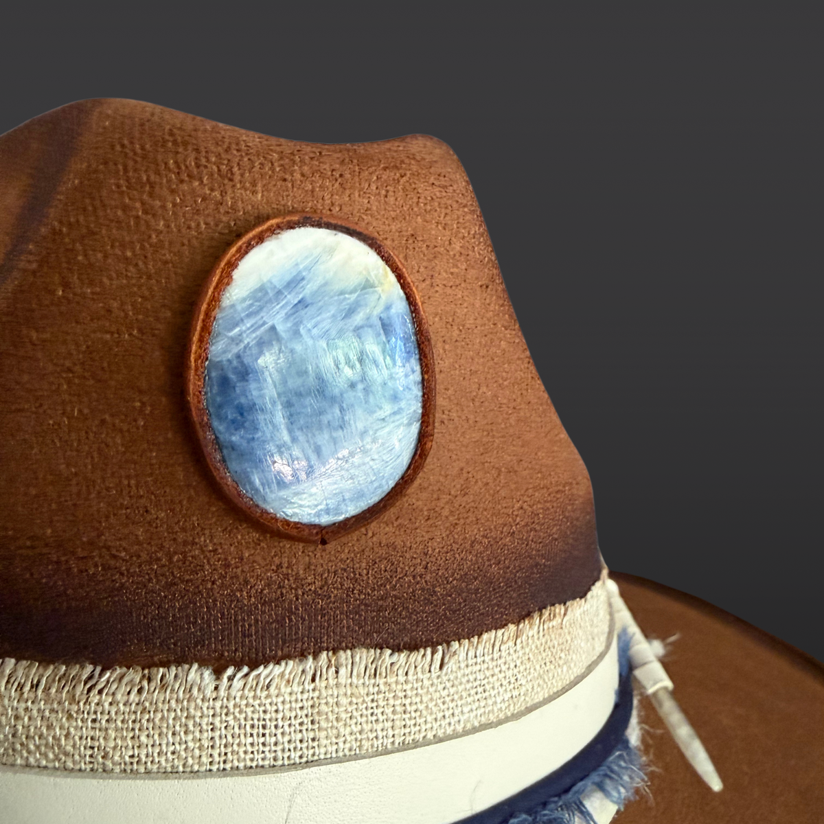 A close-up of the blue kyanite stone attached to the cream leather band of a tobacco wide-brimmed fedora.