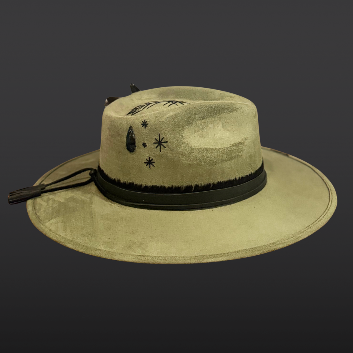 Side view: A sage green wide-brimmed fedora shown from the side, featuring a hand-embroidered arrow, black leather band with tassels, and rooster feather accents.