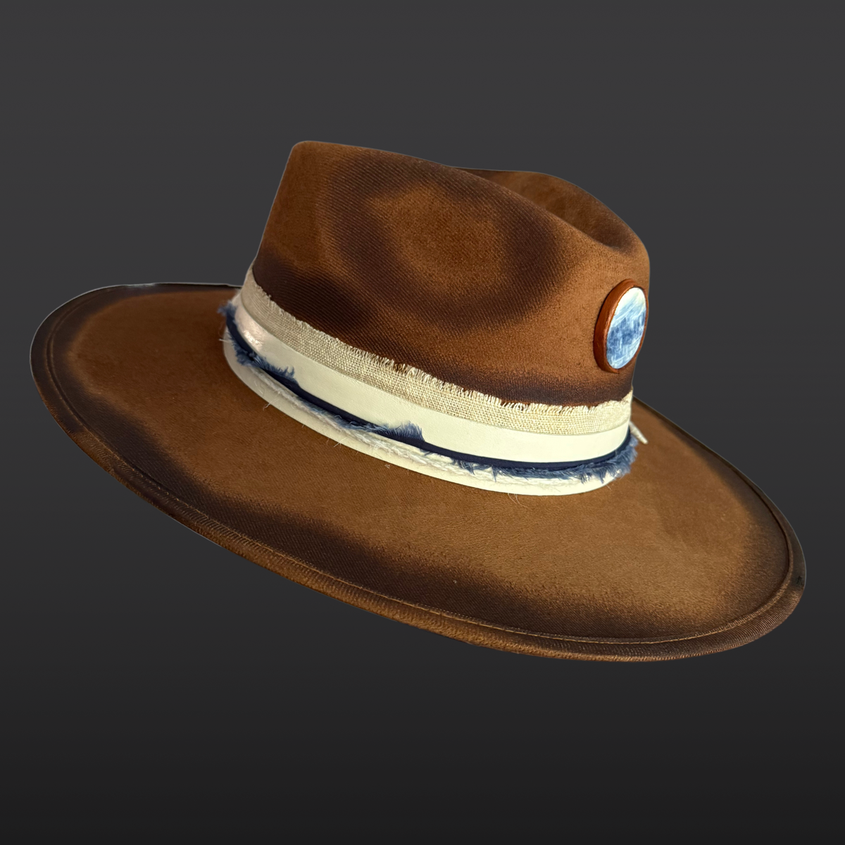 A tobacco wide-brimmed fedora shown from the right side, featuring an all-over hand-torched finish, cream leather band with navy cotton wrap, blue kyanite stone, and turkey feather accent.