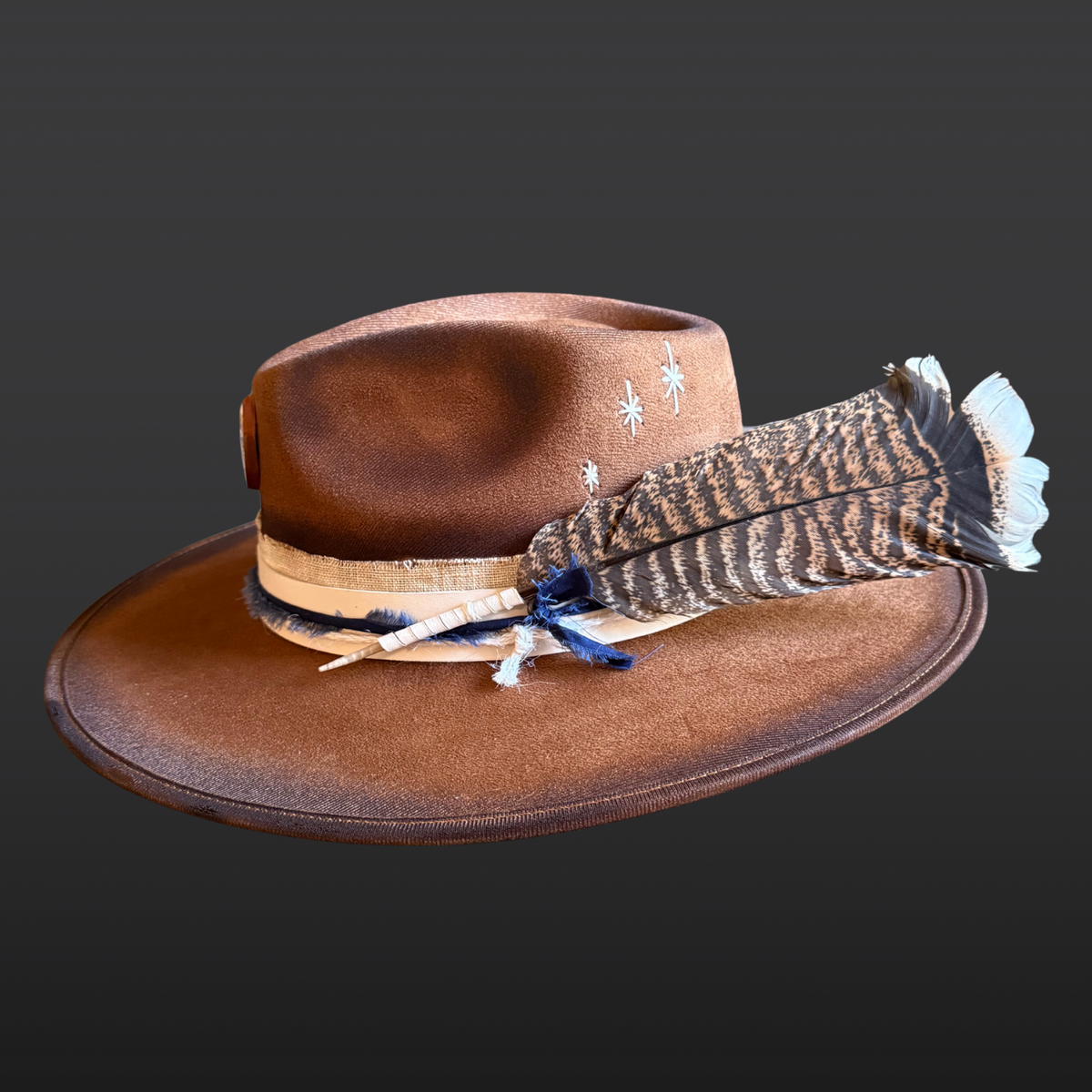 A tobacco wide-brimmed fedora shown from the left side, featuring an all-over hand-torched finish, cream leather band with navy cotton wrap, blue kyanite stone, and turkey feather accent.