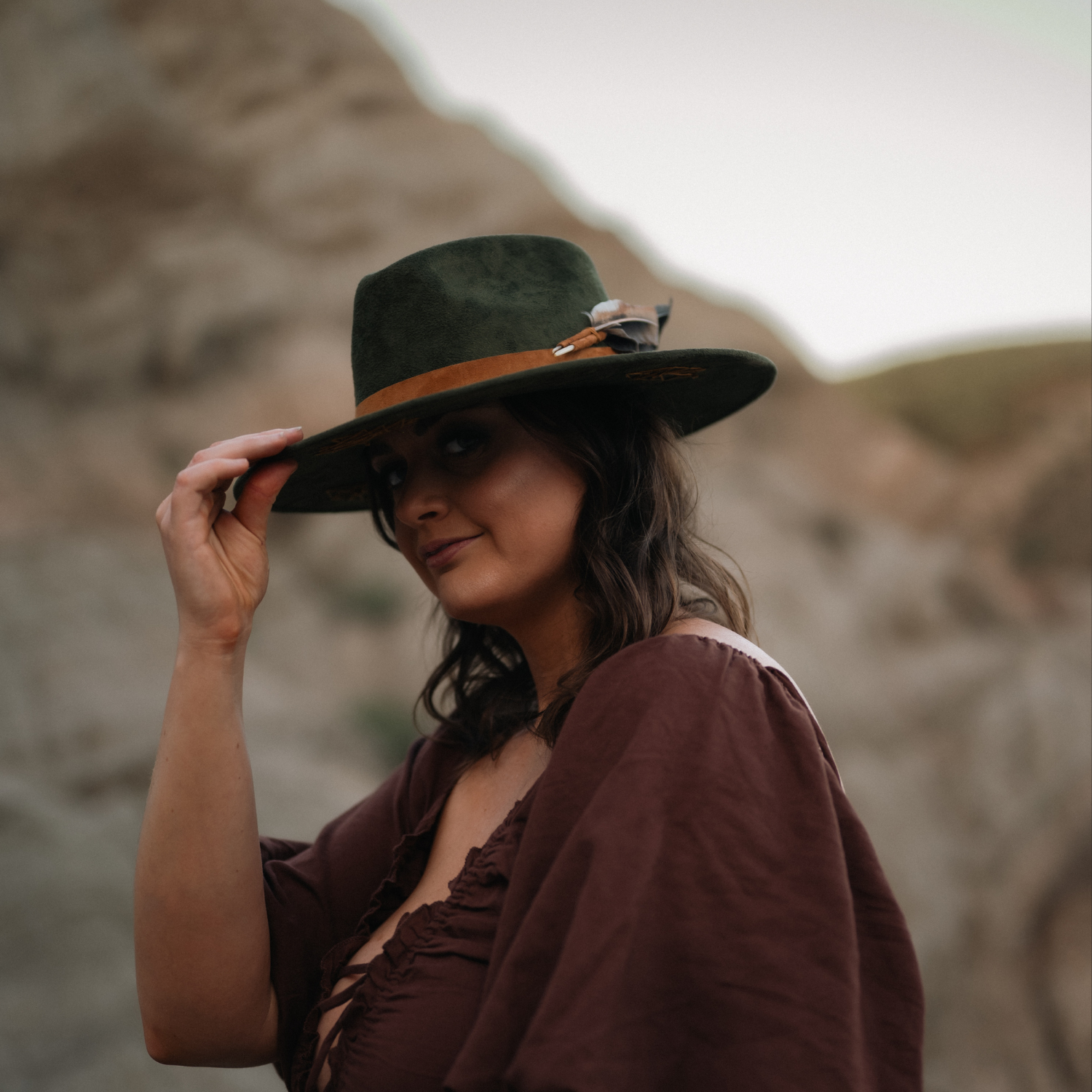 Woman in a burgundy dress wearing the Root & Radiance forest green fedora against the badlands, holding the brim