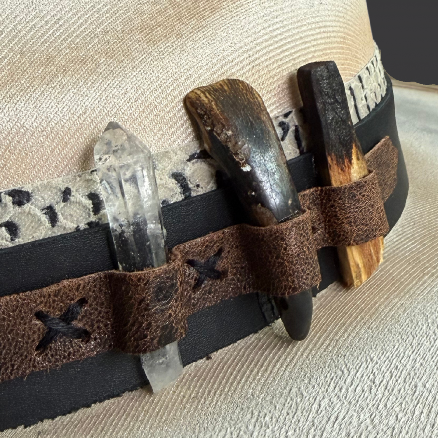 close up  details of Unbound fedora showing black and ivory snake-print leather, brown leather accent, clear quartz crystal, bison tooth, and burnt Palo Santo.