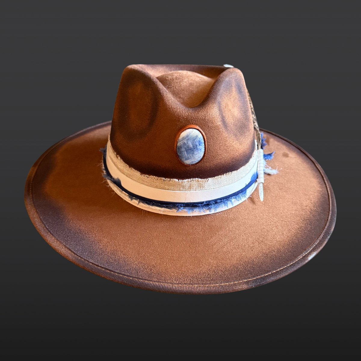 A tobacco wide-brimmed fedora featuring a hand-torched textured finish, embroidered sunburst on the crown, blue kyanite stone, and cream leather band.