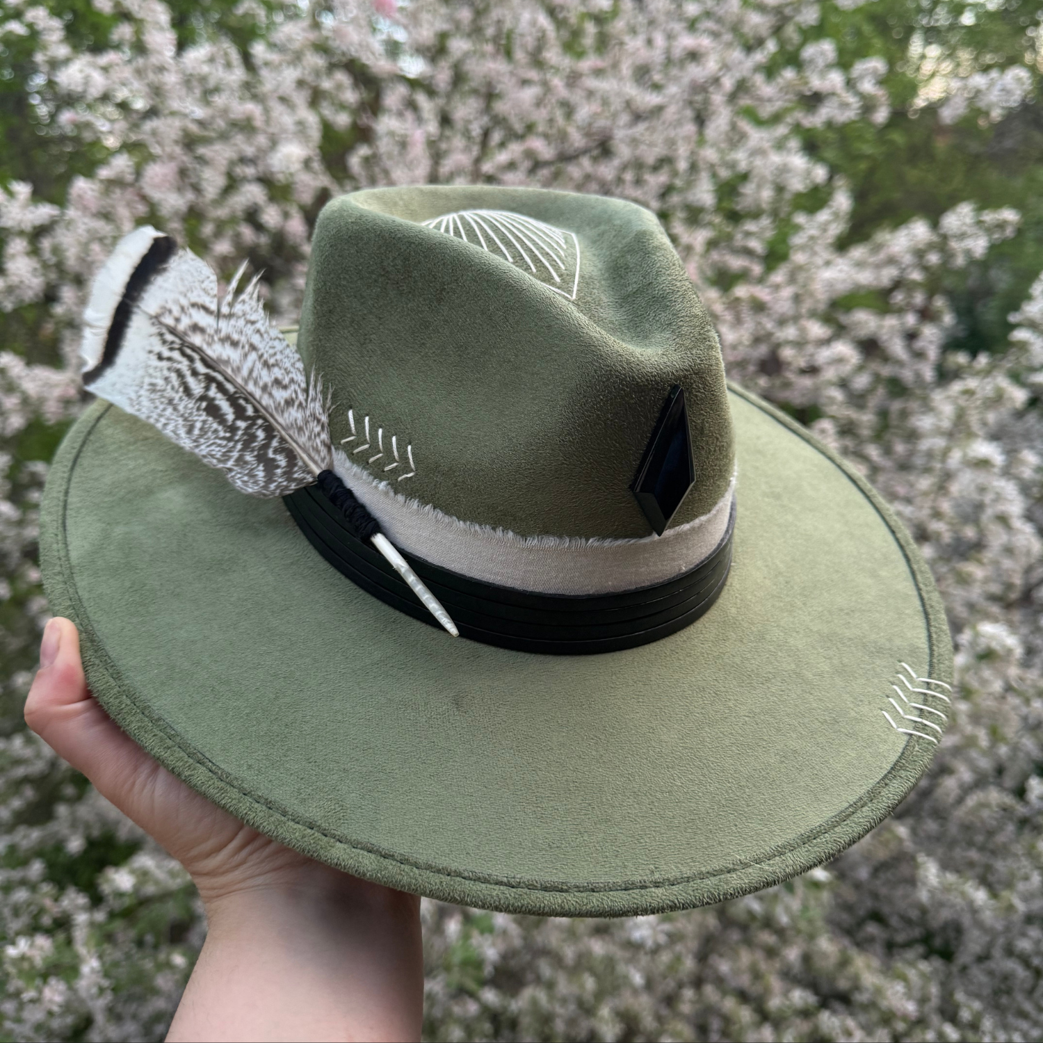 One of a kind sage green artisan fedora hat with obsidian stone, black leather band, white cotton wrap, feather detail, and hand embroidered directional symbols held infant of floral background by Inara Hat Co.