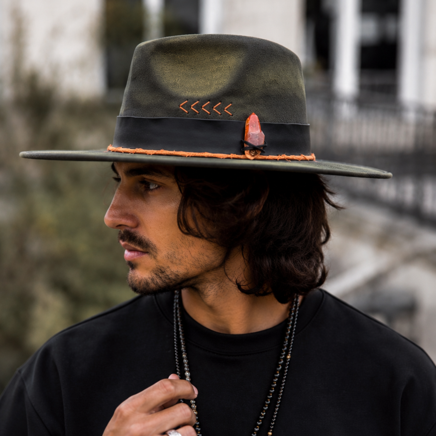 Man wearing the Solar Path Fedora Hat, a one-of-a-kind dark green wide-brim fedora featuring hand-torched vegan suede, a tangerine quartz crystal, black leather band, orange directional arrow details, and feather accents. Boho men's fashion with rugged street style and festival-inspired accessories.